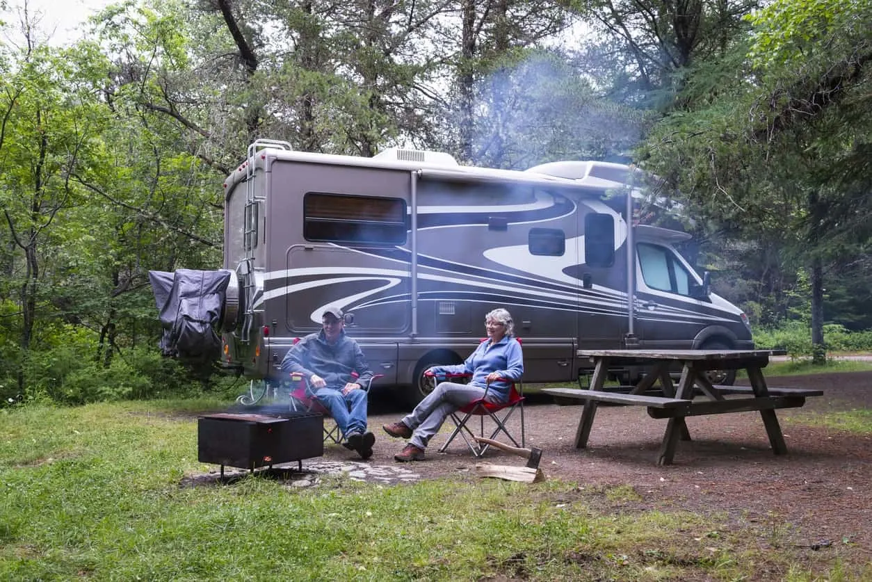 A couple sits in the middle of the woods with their campervan parked just behind them.