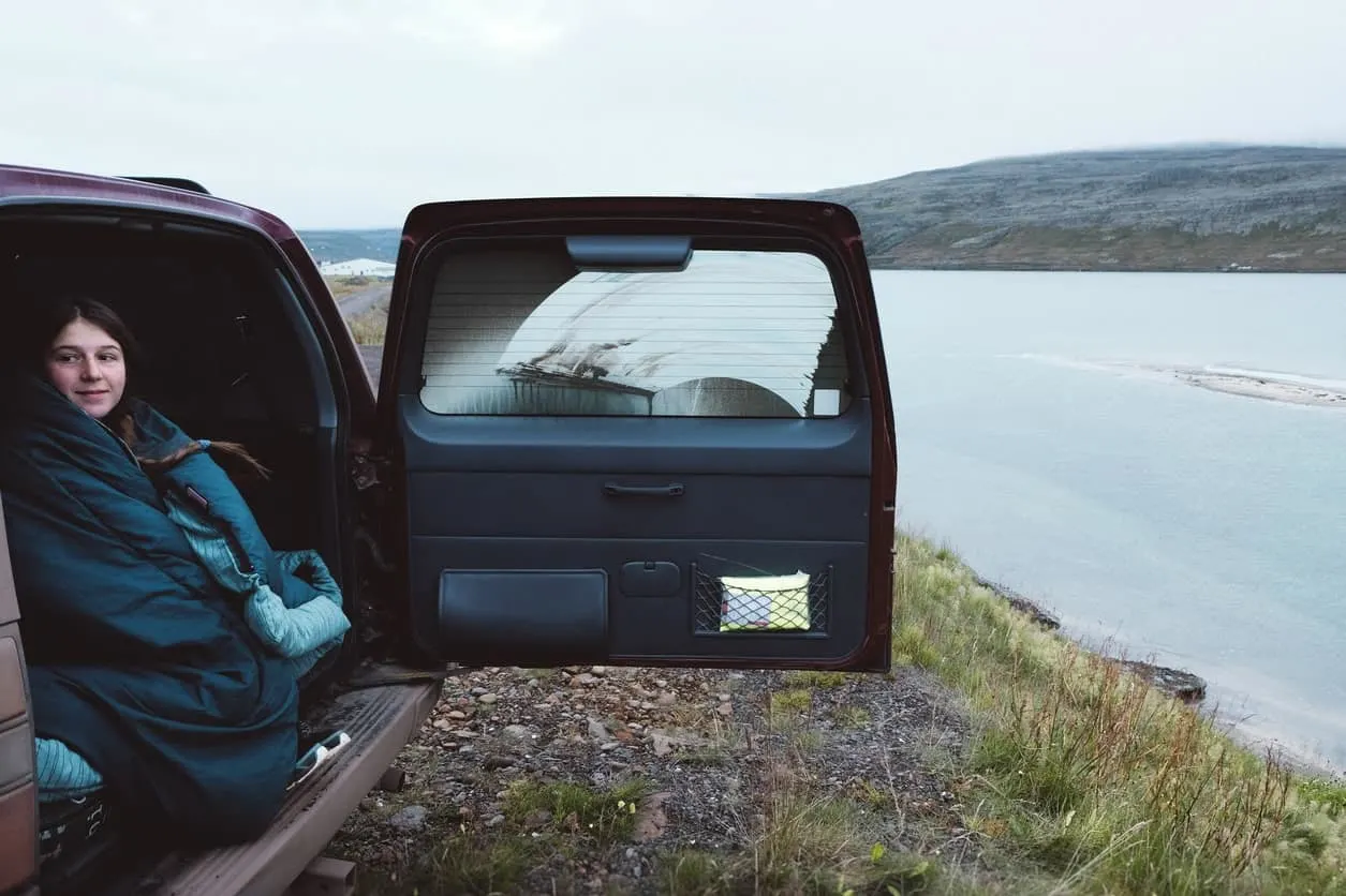 A young woman in her sleeping bag sits at the rear of a vehicle and looks out at the water.