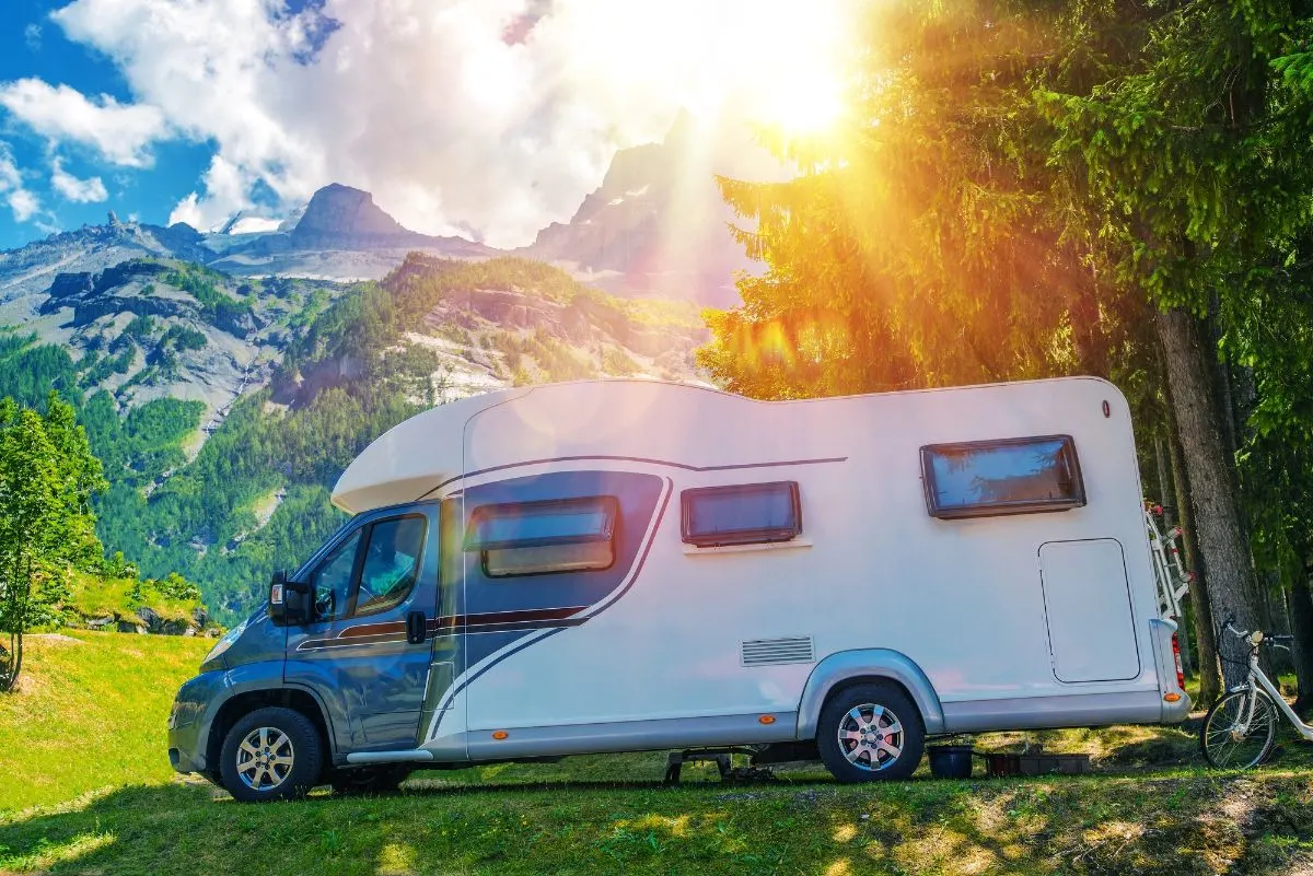 Side view of white and blue Class C Campervan on camp site with shade sun rays.