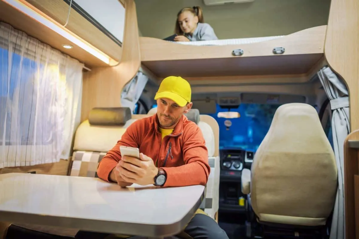 A man looks at his smartphone in the kitchenette inside an RV while a young girl is at the loft bed enjoying her gadget.