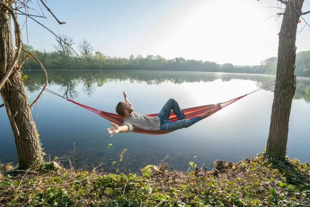 Man extends his arms out as he lies on a hammock between trees and in full view of the lake.