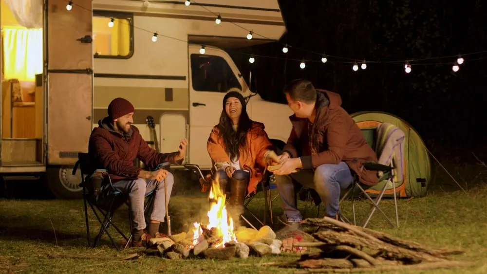 A group of friends around a camp fire with the RV and the tent behind them.