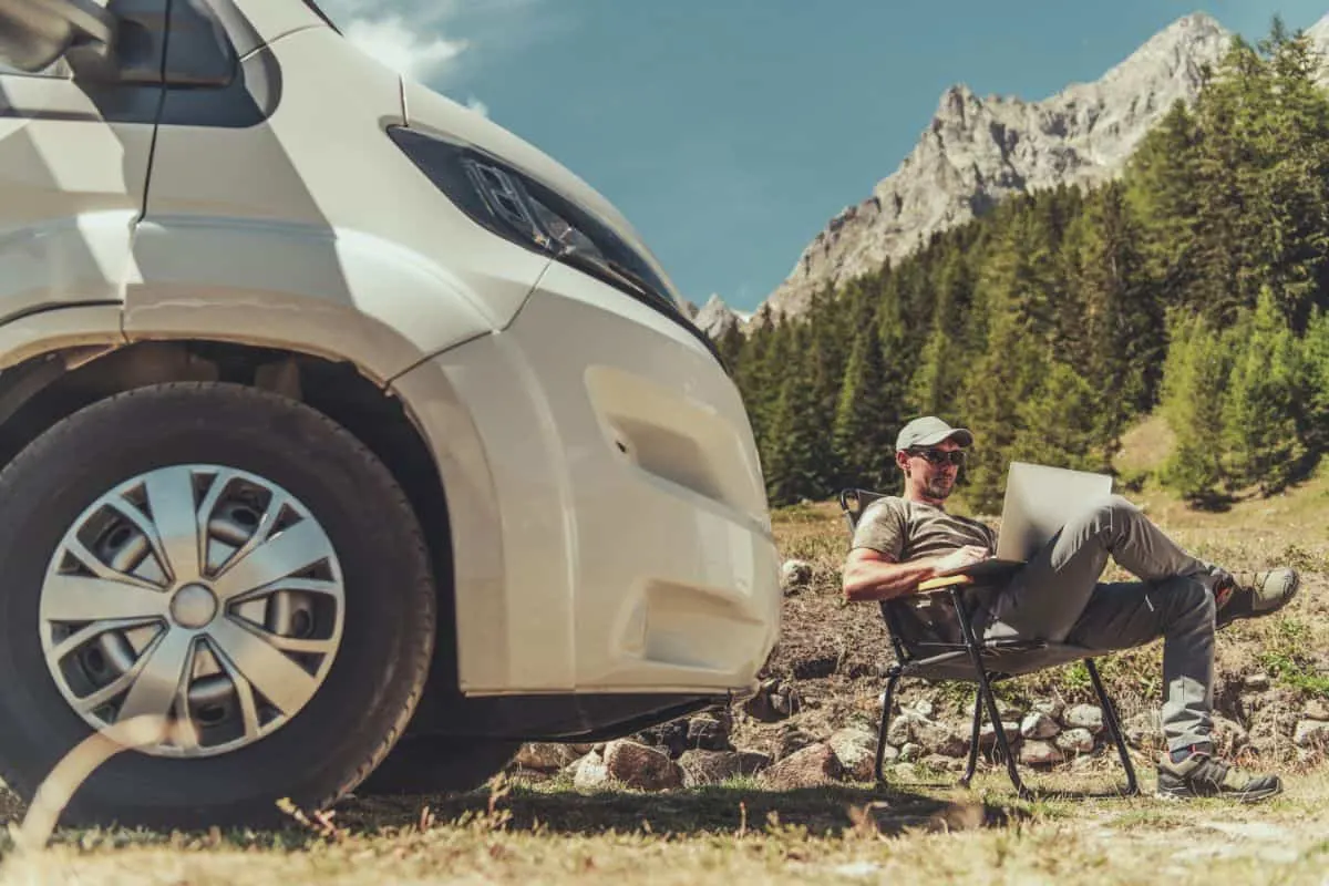 A man works on his laptop beside a parked RV outdoors.