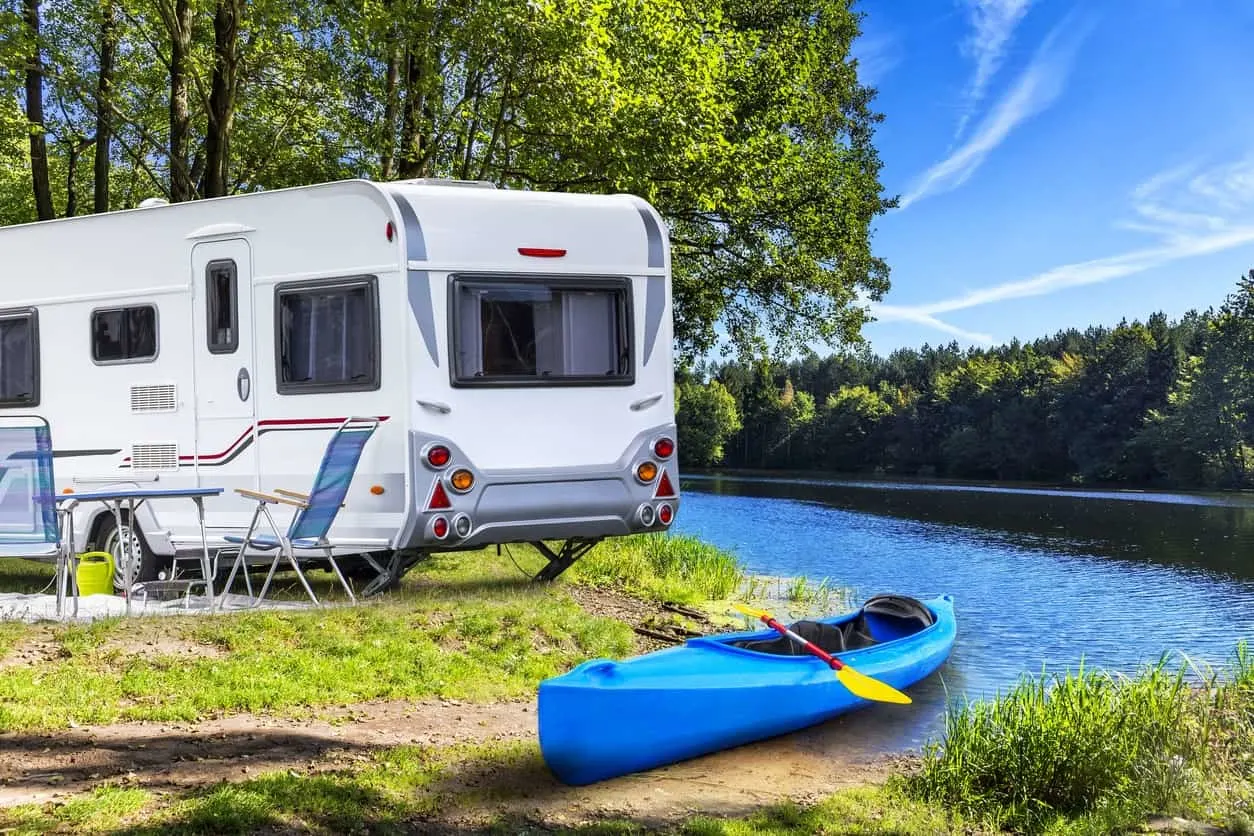 An RV trailer is parked beside a table and chairs while a canoe is docked at the edge of the lake.