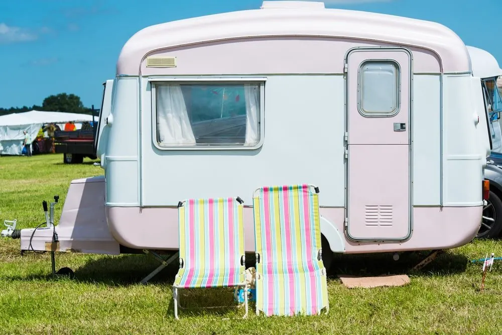 A small travel trailer with white and pink pastel tones that match the outdoor chairs.