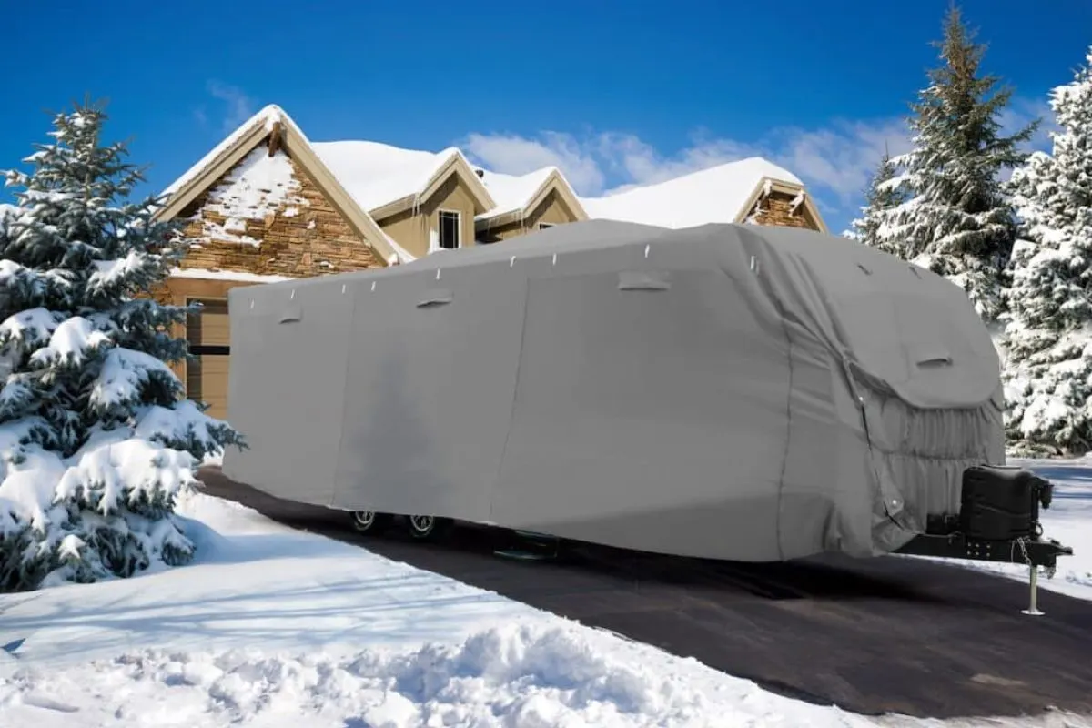 An RV with cover parked outside someones house driveway in the snowy winter.