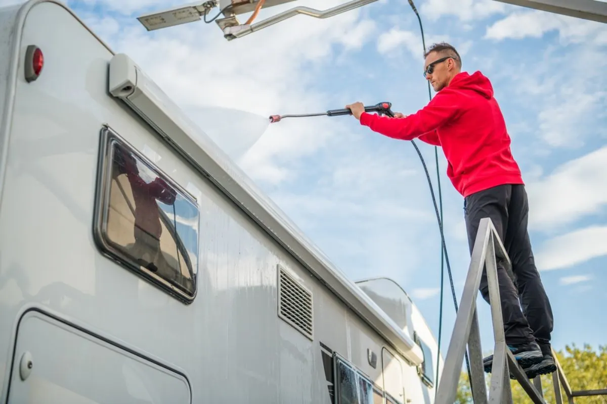 A man wearing a red jacket washing his rv using a water hose.