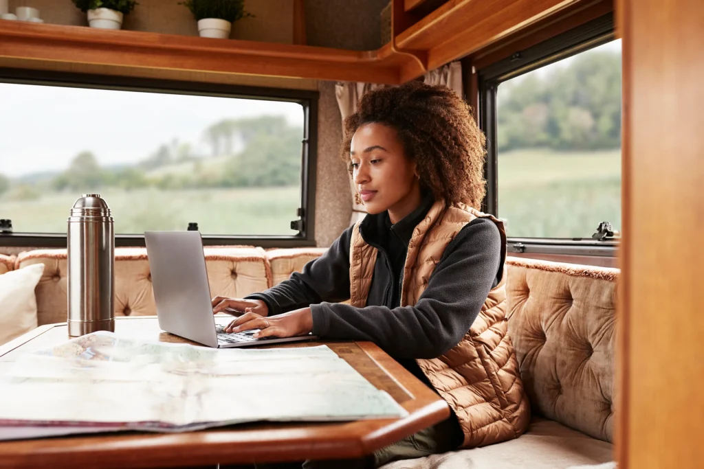 A woman working on a laptop in a RV with a field and forest on a background