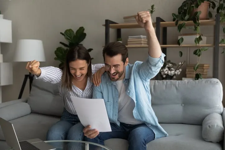 A couple sitting on a couch in their living room, smiling and cheering with their fists raised in excitement while looking at a paper they are holding, possibly celebrating good news.