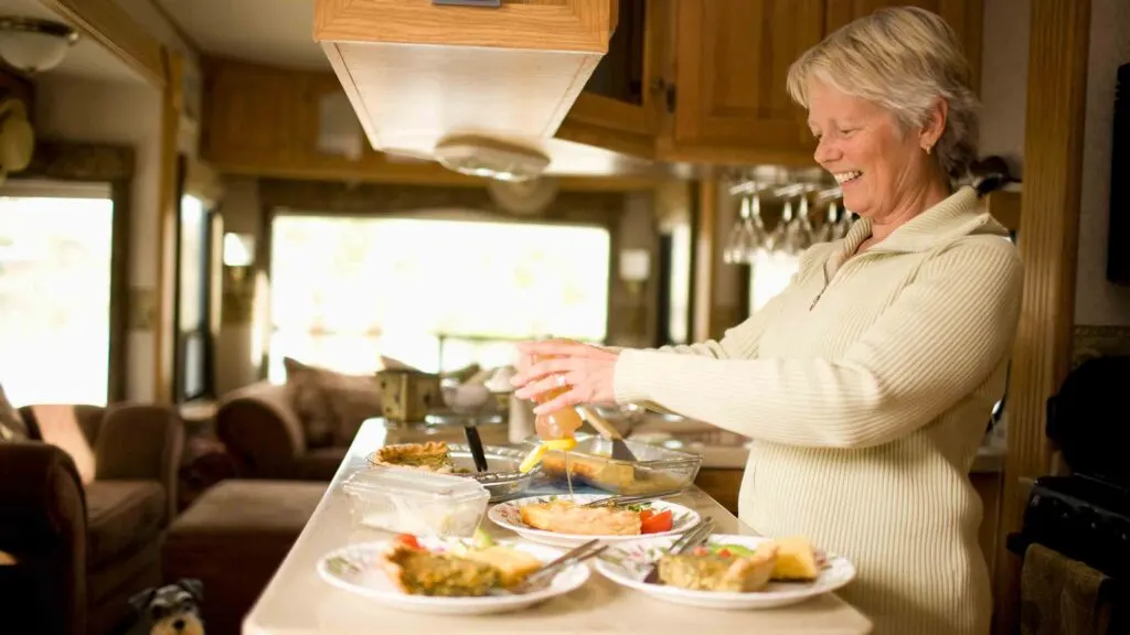 A smiling older woman preparing food in the kitchen area of a motorhome, with various dishes and ingredients on the counter.