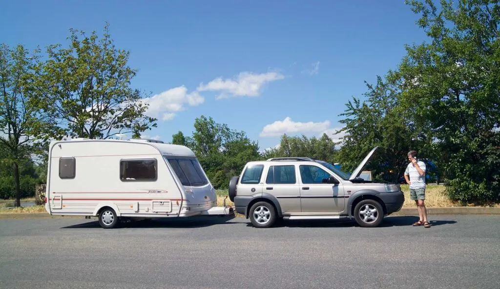 An SUV with its hood open connected to a travel trailer, with a man standing nearby talking on a phone.