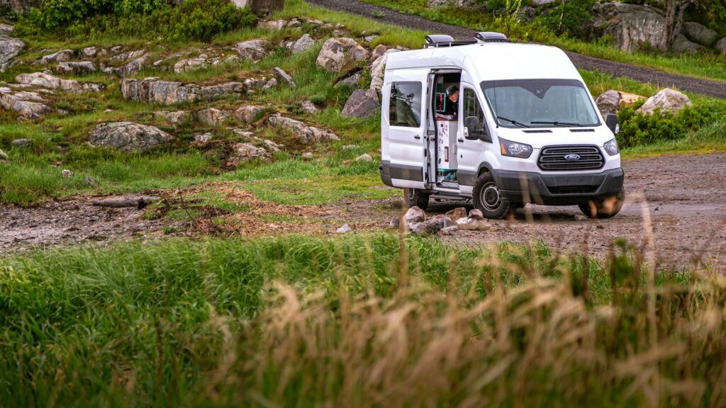  A white campervan with its door open, parked on a rocky and grassy area with greenery in the background. A person is partially visible inside the van.