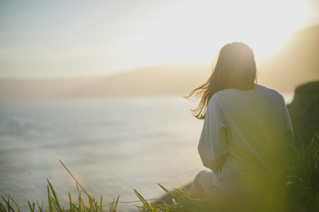  A person sitting on a grassy cliff overlooking the ocean, their back to the camera, with the sun shining brightly in the sky.