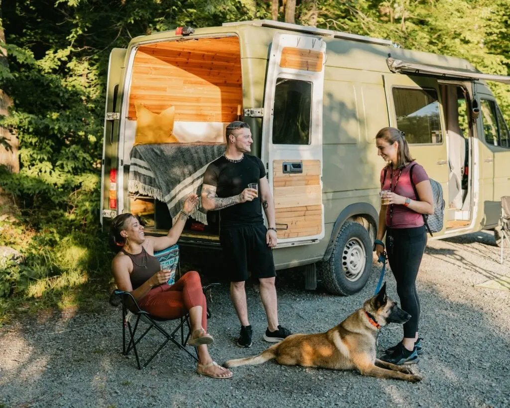 Three people and a dog relaxing outside a camper van, with wooded surroundings and sunlight casting shadows.