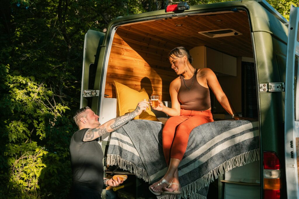 A couple enjoying a drink, with one sitting inside a cozy, wood-lined camper van and the other standing outside, in a forested location.