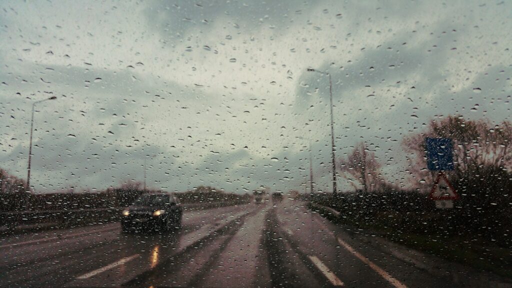 A view of a rainy highway through a windshield with raindrops on the glass and blurred vehicles in the background.