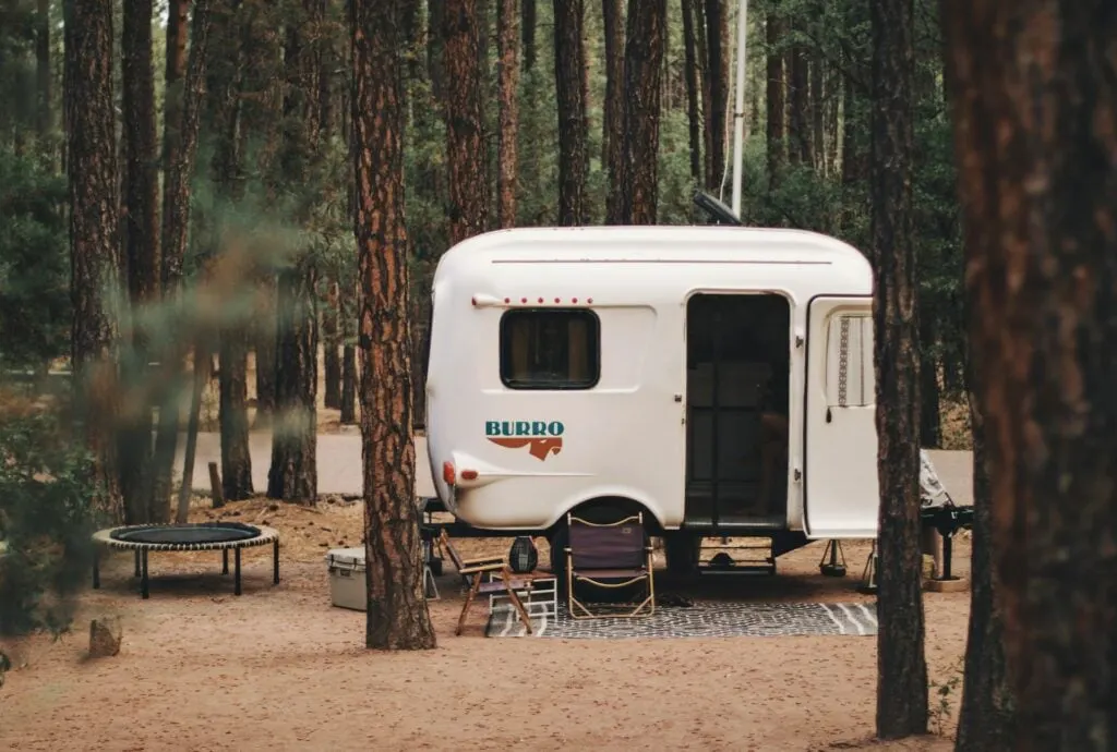 A small white camper labeled "BURRO" parked in a forest, with camping chairs and a trampoline outside.