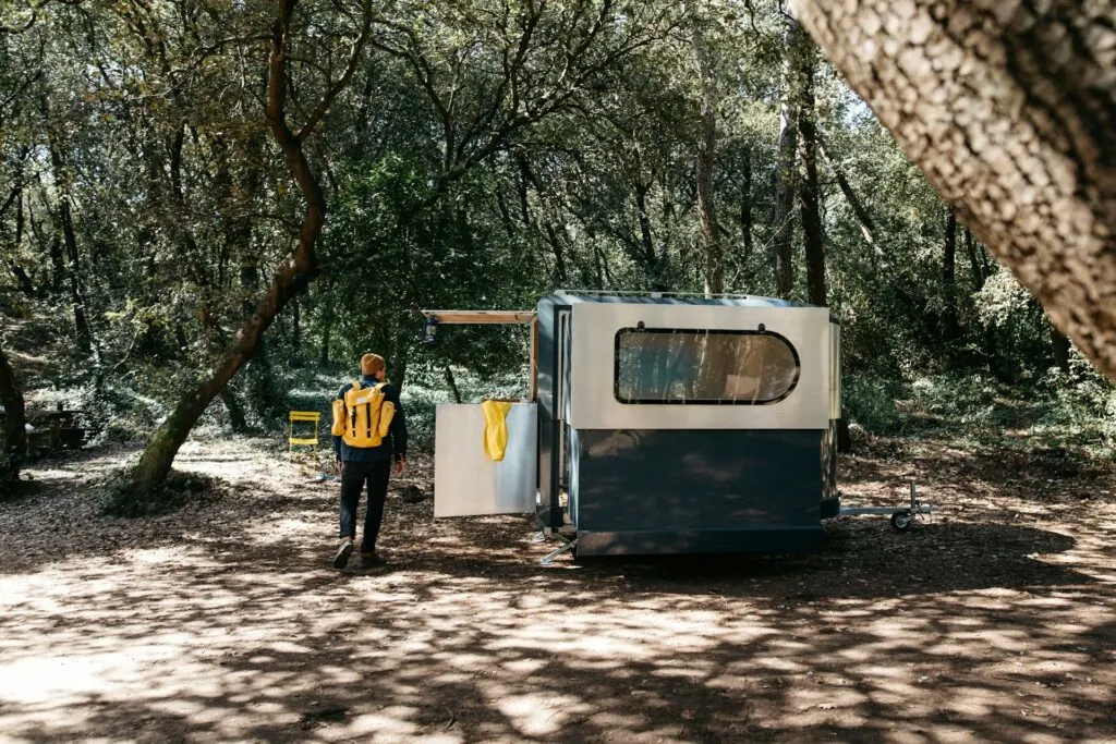 A person wearing a yellow backpack and beanie stands next to a compact camper trailer in a wooded area.