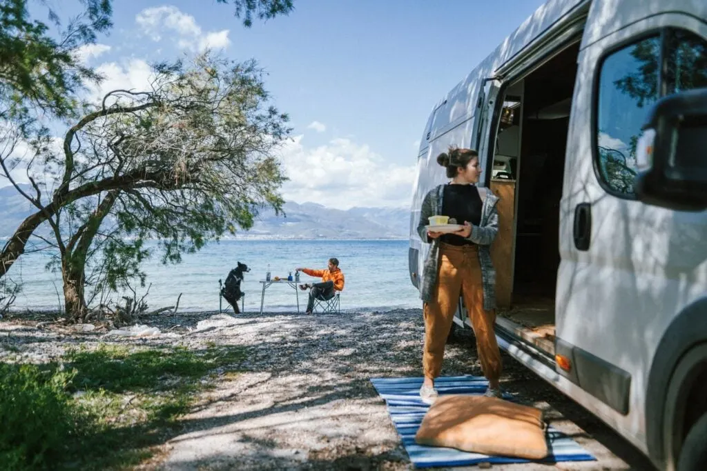 A woman standing near an RV parked beside a shoreline, holding a plate and looking at another person sitting at a table with a dog under a tree. The scenic background includes mountains, water, and a clear sky.