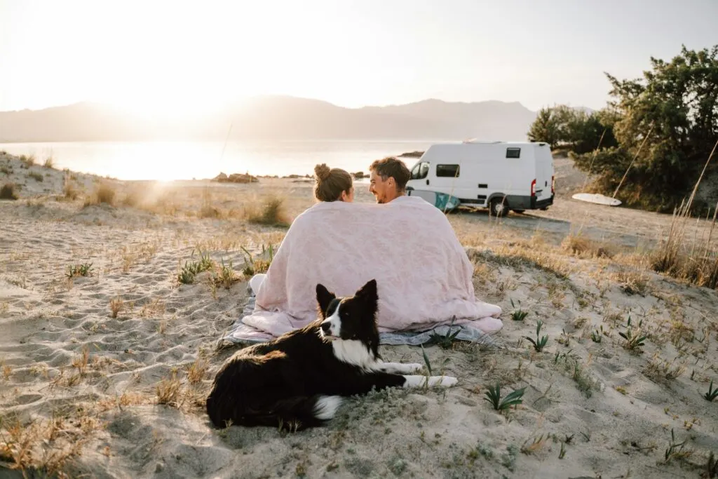 A couple sitting on a sandy beach wrapped in a pink blanket, watching the sunset over a body of water with mountains in the background. Their dog lies nearby, and a camper van is parked in the distance.