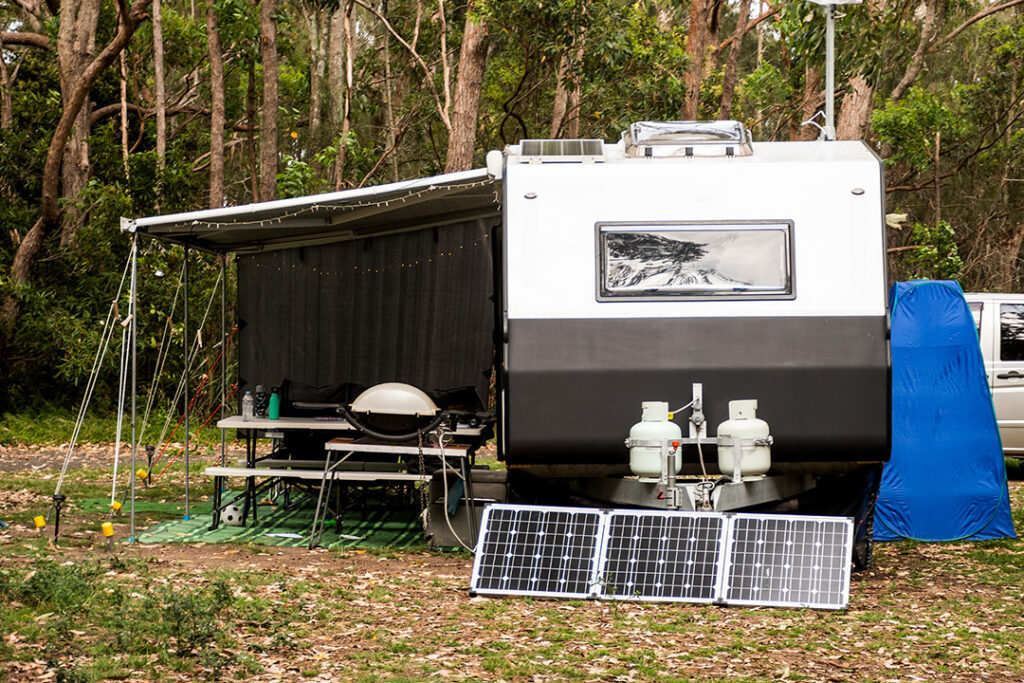 A camping setup with a modern RV featuring outdoor furniture, a grill, and solar panels positioned at the front, in a wooded area.