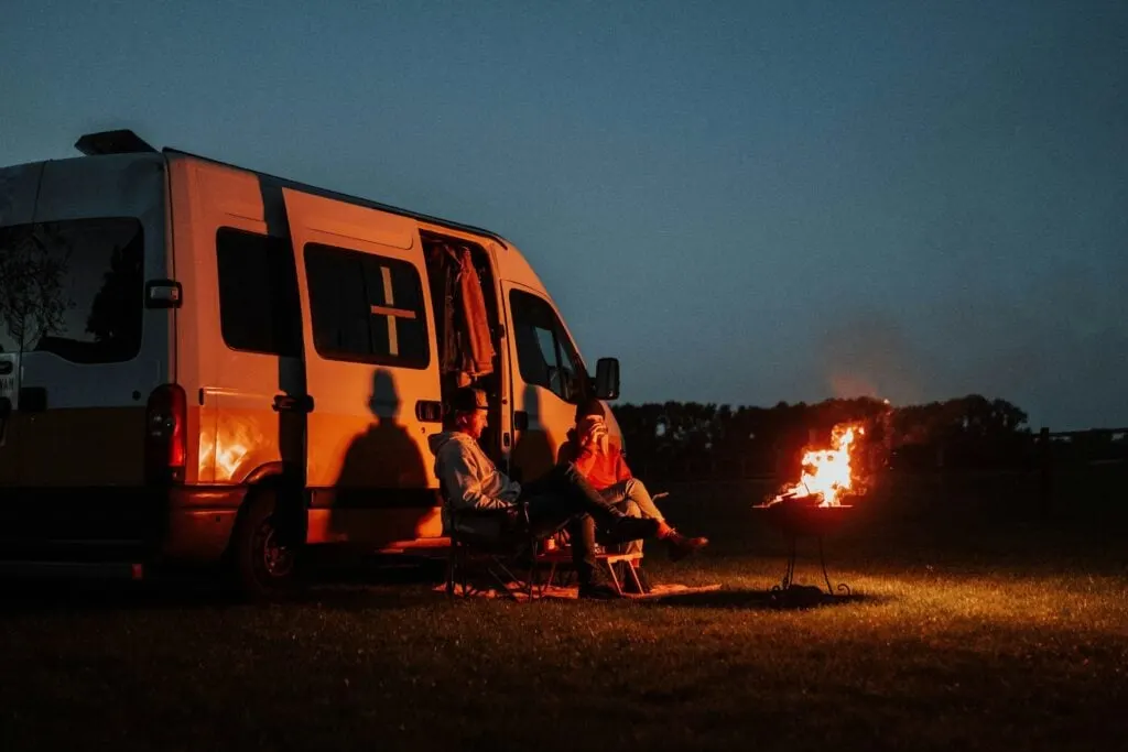 Two people sitting in chairs next to a van, warming themselves by a campfire at night.