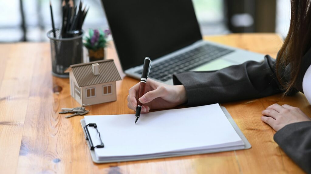 A person signing documents on a clipboard at a wooden desk with a laptop, a small model house, keys, and a pencil holder in the background.