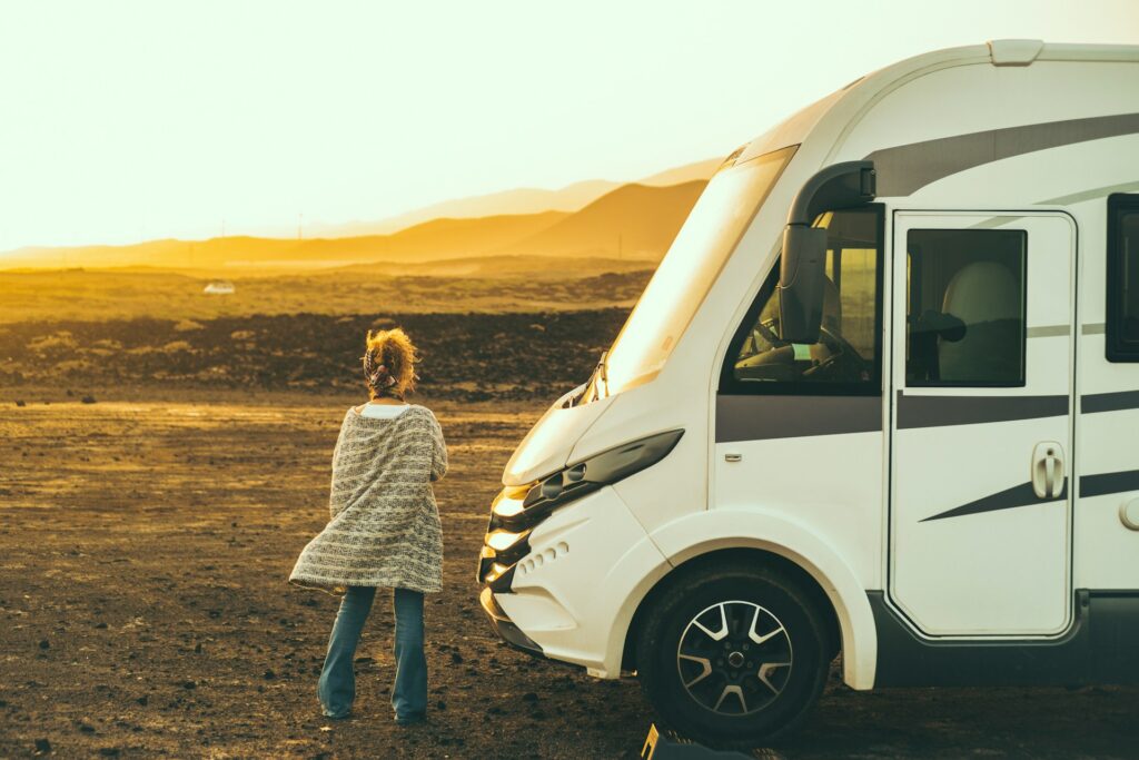 A person standing beside a white campervan, looking out at a barren landscape during sunset with mountains in the distance.