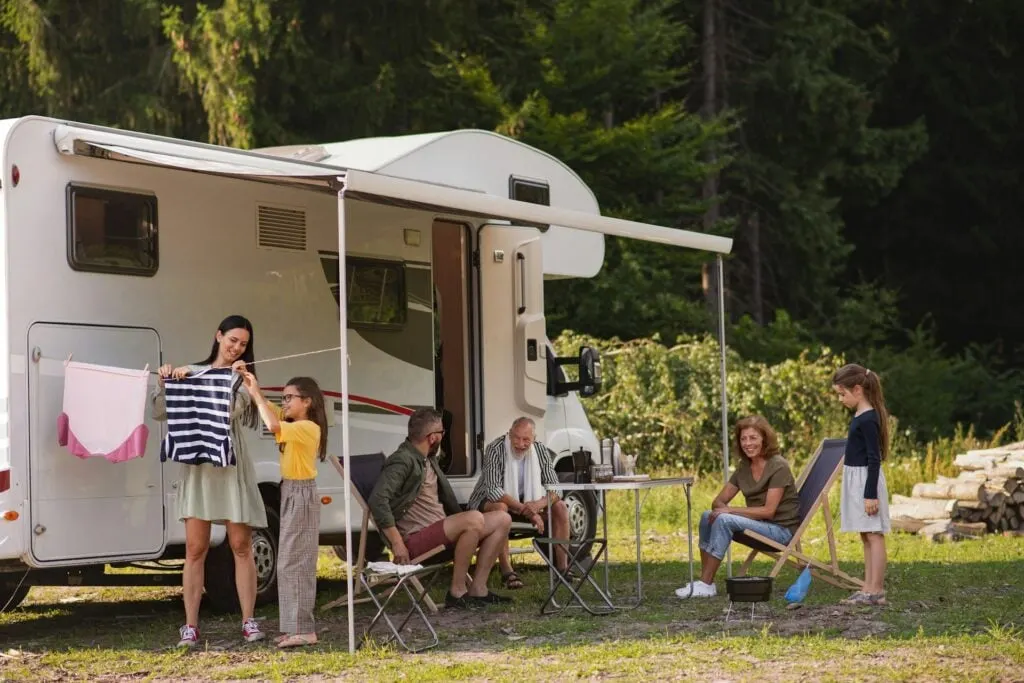 A family enjoying a camping trip outside their motorhome. Two people hang laundry on a line, while others sit at a table or on a chair, surrounded by trees.