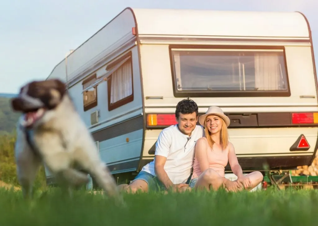 A couple sitting on grass near a camper with a dog running in the foreground, enjoying a sunny day outdoors.