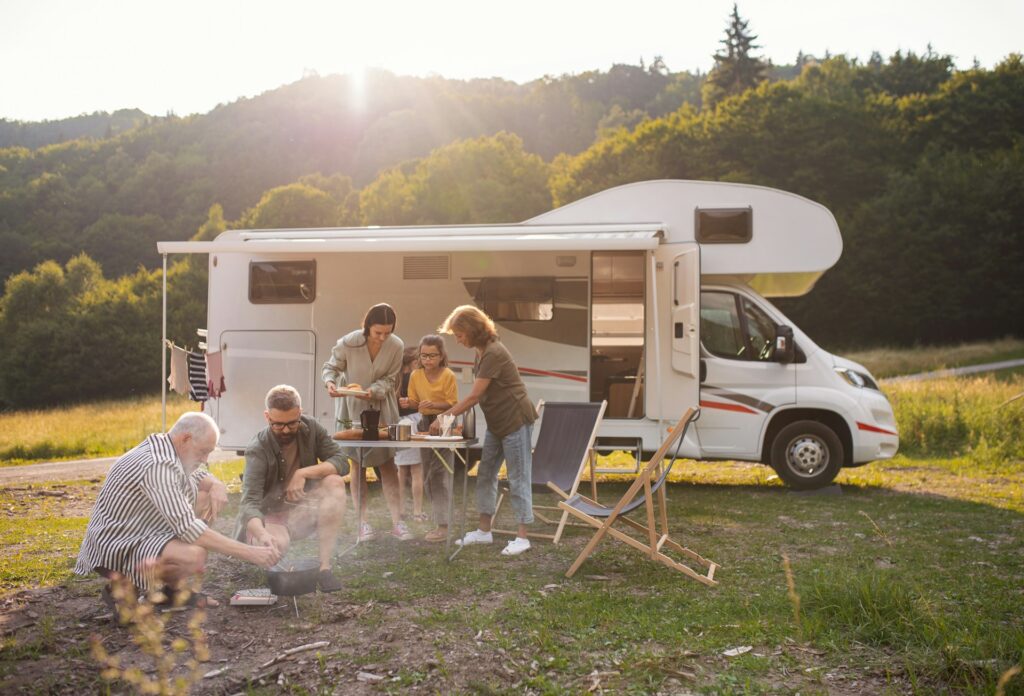 A group of people gathered around a table and campfire next to an RV, surrounded by nature, with the sun setting behind the hills in the background.