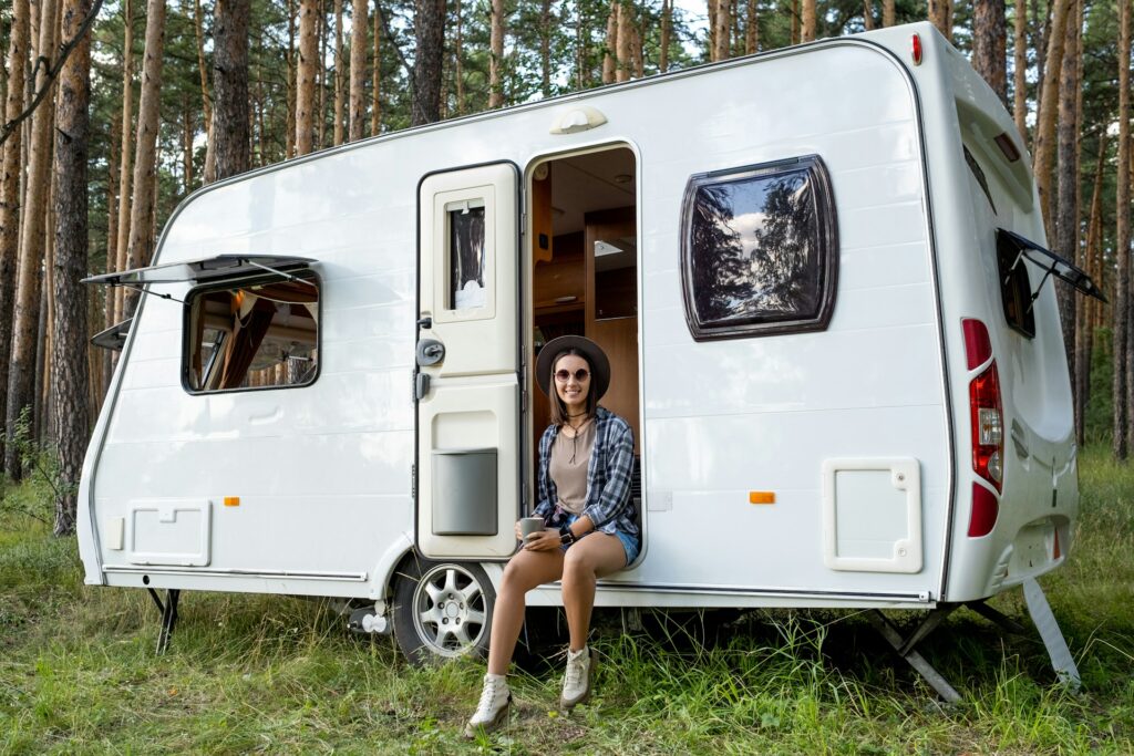 A woman sitting at the entrance of a white travel trailer in a forested area.