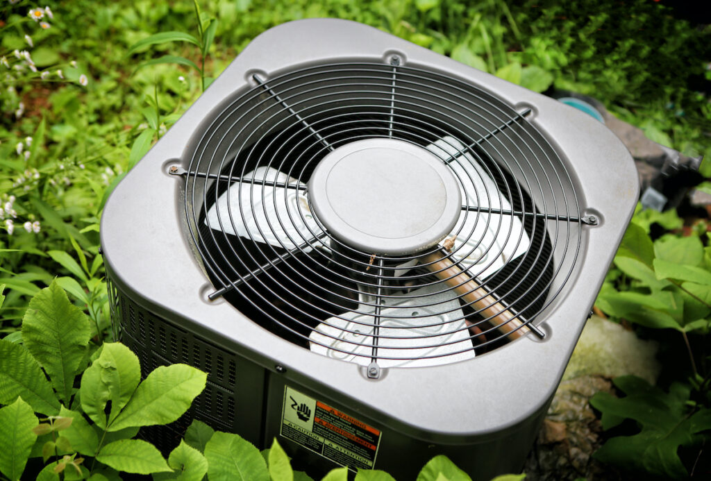  A close-up of an air conditioning unit's outdoor component surrounded by green plants.