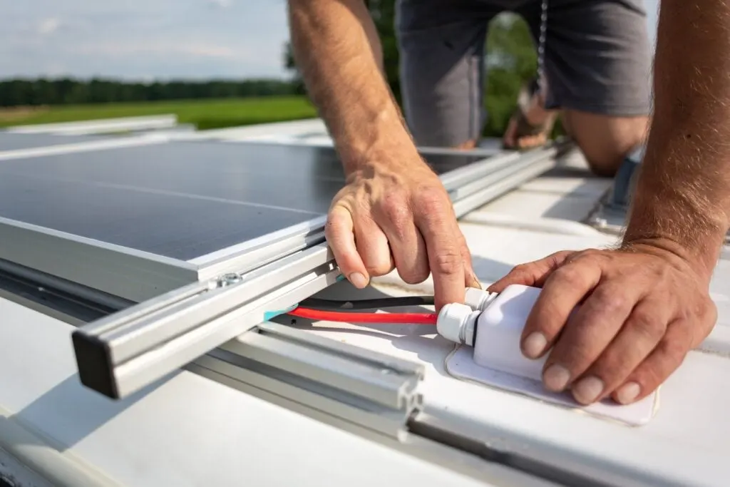 A close-up of a man's hands connecting wires to a solar panel mounted on the roof of an RV.