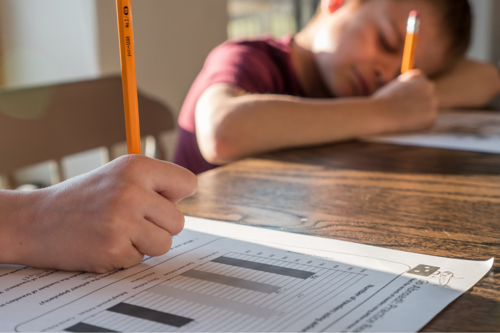 Close-up of a student's hand holding a pencil and working on a worksheet at a wooden table, with another student visible in the background.