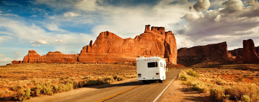 A motorhome travels down a road flanked by towering red rock formations under a partly cloudy sky.