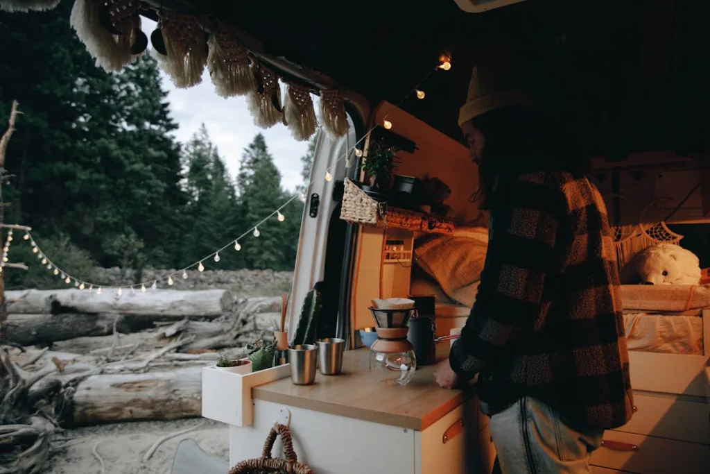 A man standing inside a cozy camper van setting up a drink, with the back doors open, revealing an outdoor scene with string lights and trees.