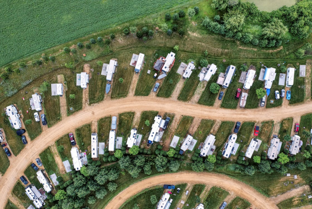 An aerial view of an orderly RV park with rows of RVs and vehicles on grassy plots, bordered by trees.