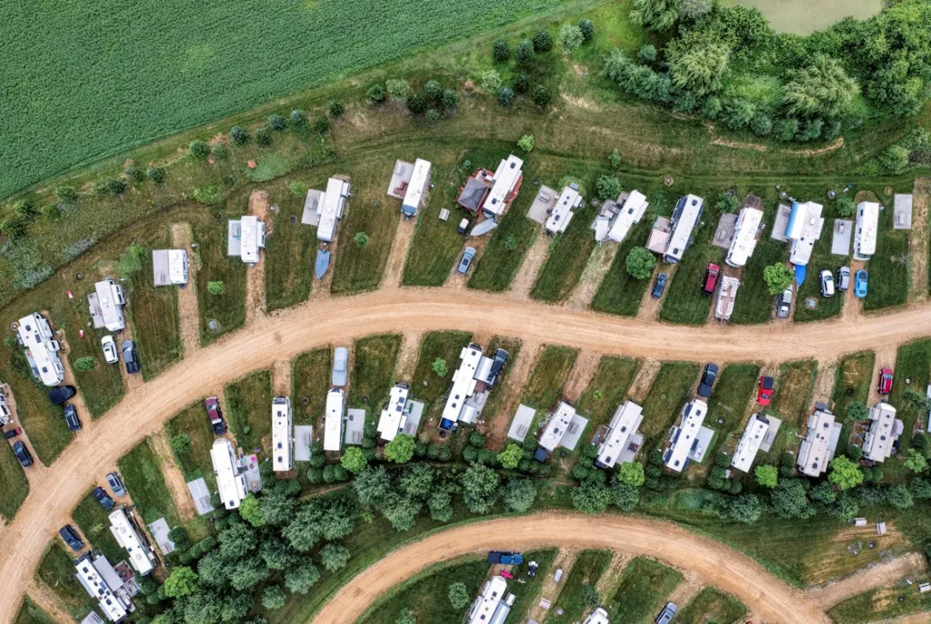 An aerial view of an orderly RV park with rows of RVs and vehicles on grassy plots, bordered by trees.