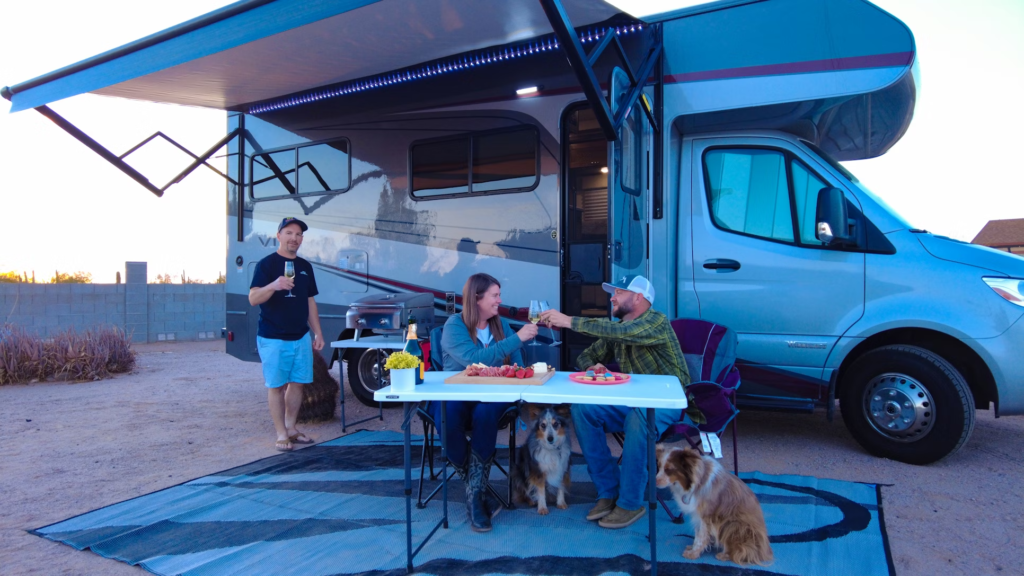 A group of people enjoying a meal and drinks outside a motorhome, with two dogs sitting nearby.