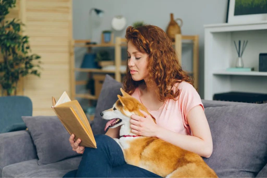 A woman with curly hair sitting on a couch, reading a book while her dog sits on her lap, in a cozy living room environment.