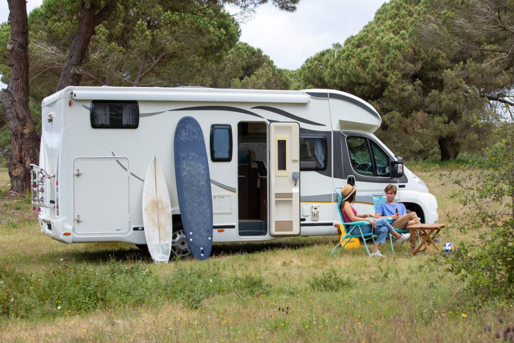 A white campervan parked on a grassy area with surfboards leaning against it. Two people are sitting on folding chairs beside a small table, enjoying the outdoors.