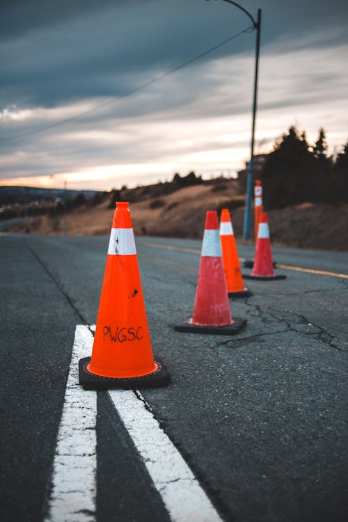 Several orange traffic cones with white reflective stripes arranged on a road, with a sunset sky and landscape in the background.