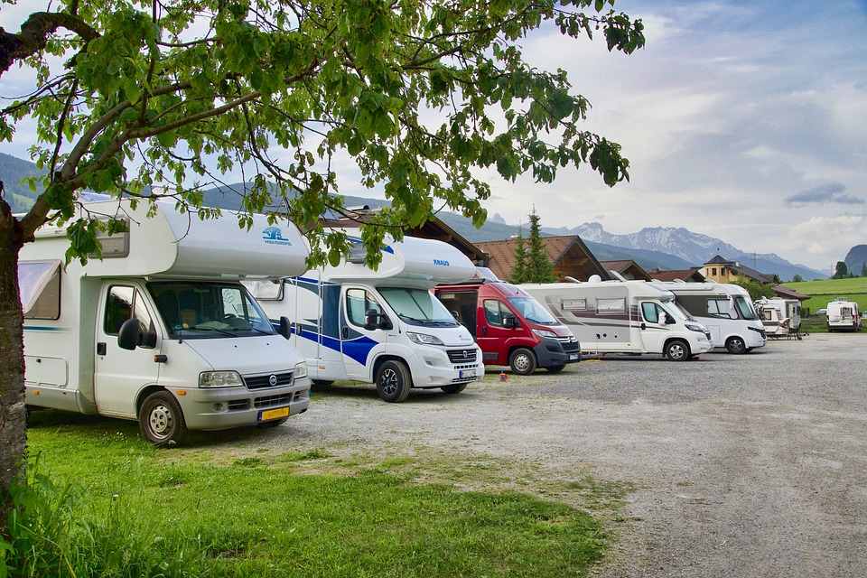 Several motorhomes parked in a row at a campsite with mountains and houses in the background, and a tree with green leaves in the foreground.