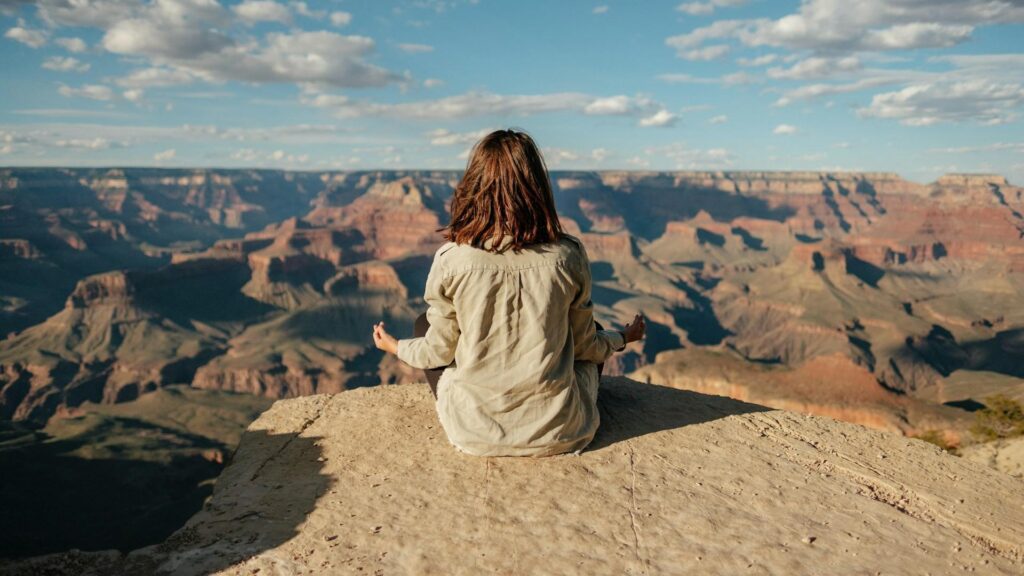 A woman sitting on a rocky edge, meditating, overlooking the vast expanse of a canyon under a partly cloudy sky.