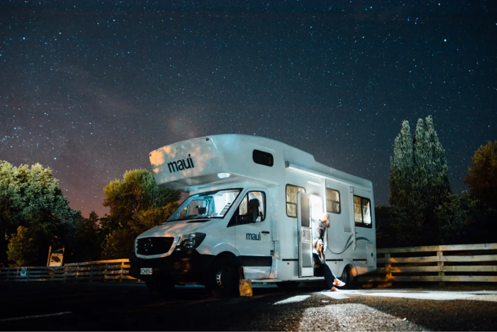 An RV with "maui" branding parked under a starry night sky, a person standing at its open door.