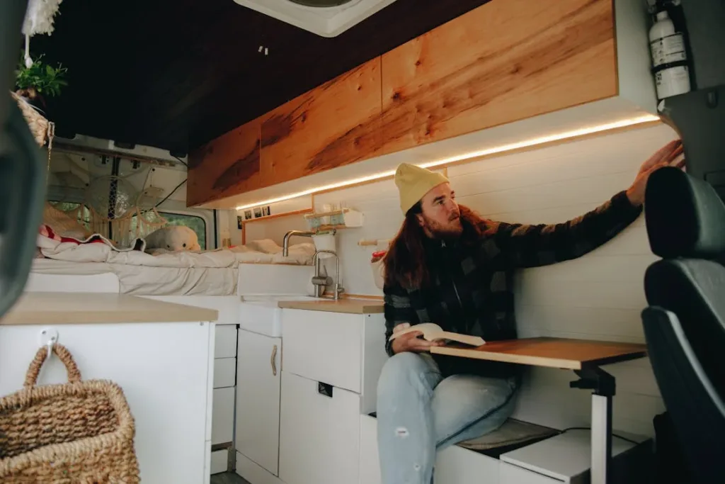 A man reading a book while sitting inside a camper van, with a neatly organized interior including a bed, shelves, and wooden cabinetry.