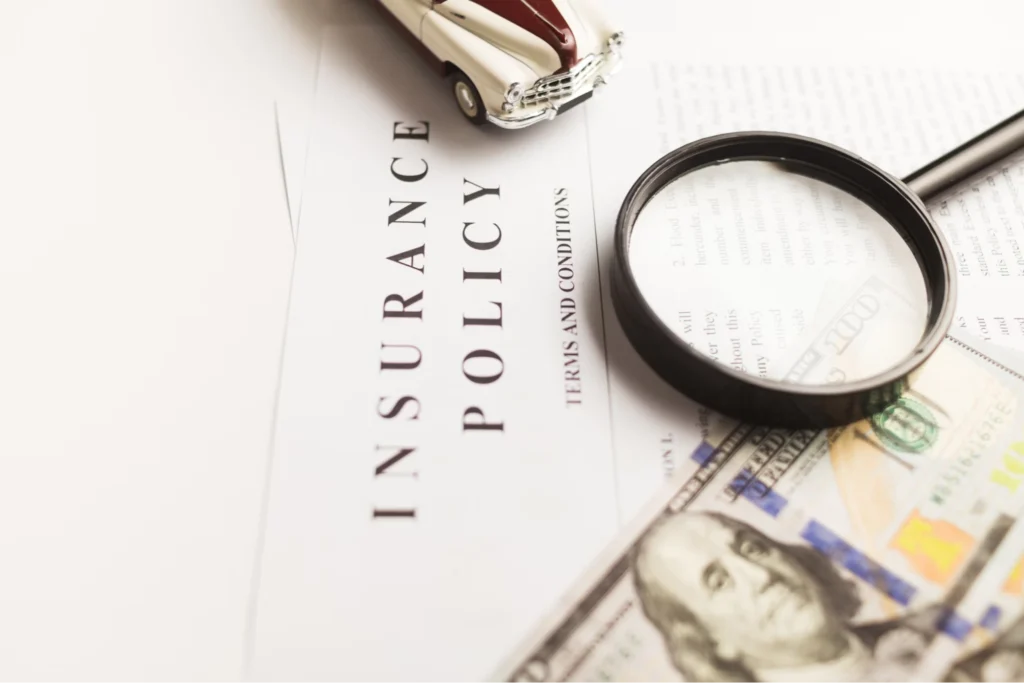 An insurance policy document with a magnifying glass placed over it, a hundred-dollar bill, and a miniature toy car on top.