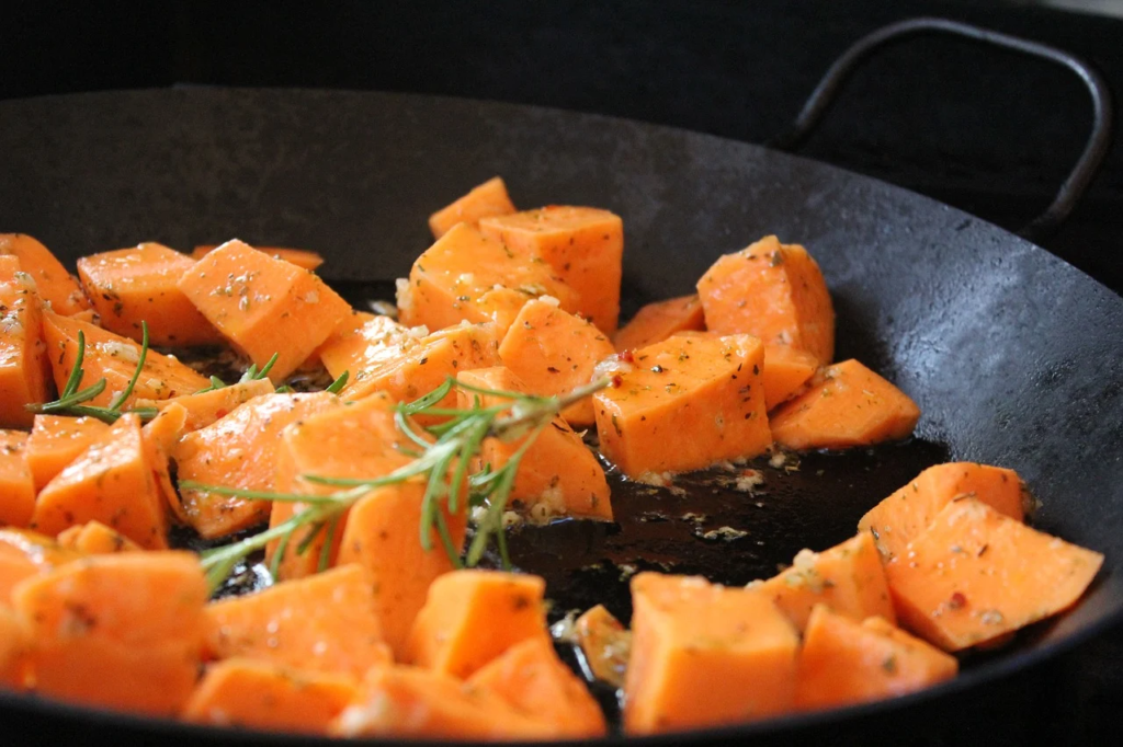 Cubes of seasoned orange vegetables, possibly sweet potatoes, with herbs, roasting in a black pan.