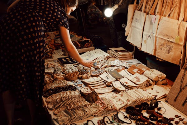 A woman in a printed dress looking at items displayed on a table at a market, with various handmade goods and bags.